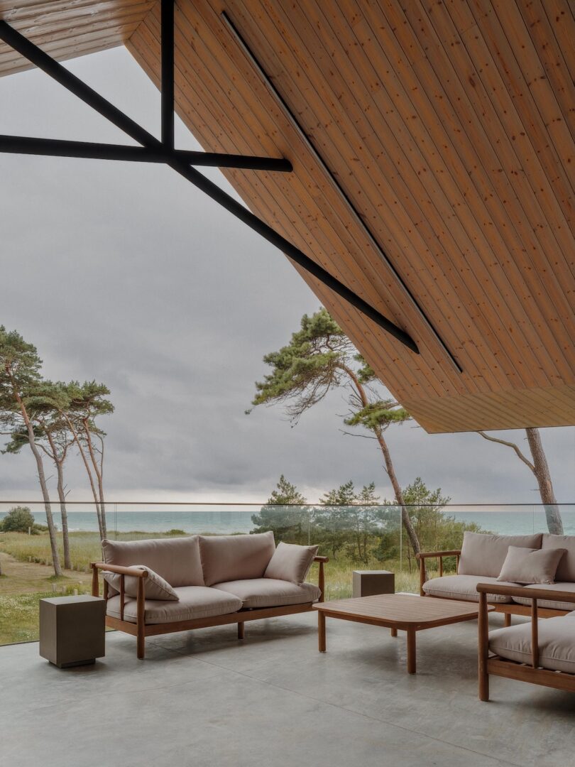 Modern outdoor patio with wooden ceiling, beige cushioned sofas, a coffee table, and a glass railing overlooking grassy landscape and sparse trees under a cloudy sky.