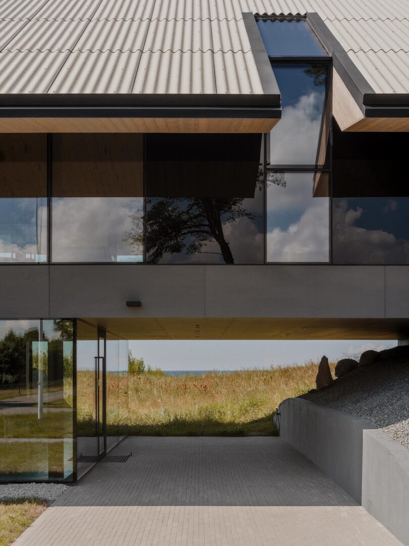 Modern house with large glass windows reflecting trees and sky, elevated above a paved entrance, surrounded by grass and gravel landscaping.