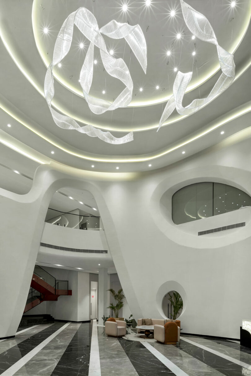 Modern lobby with curved white walls, black and white marble flooring, circular ceiling lights, and abstract hanging sculptures creates a confluence of art and design; minimalist seating features tan chairs and small tables.
