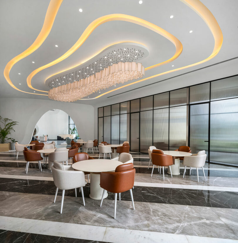 Modern lounge area with round tables, tan and brown chairs, large decorative ceiling light, marble floors, and a glass partition wall creates a confluence of style and comfort.