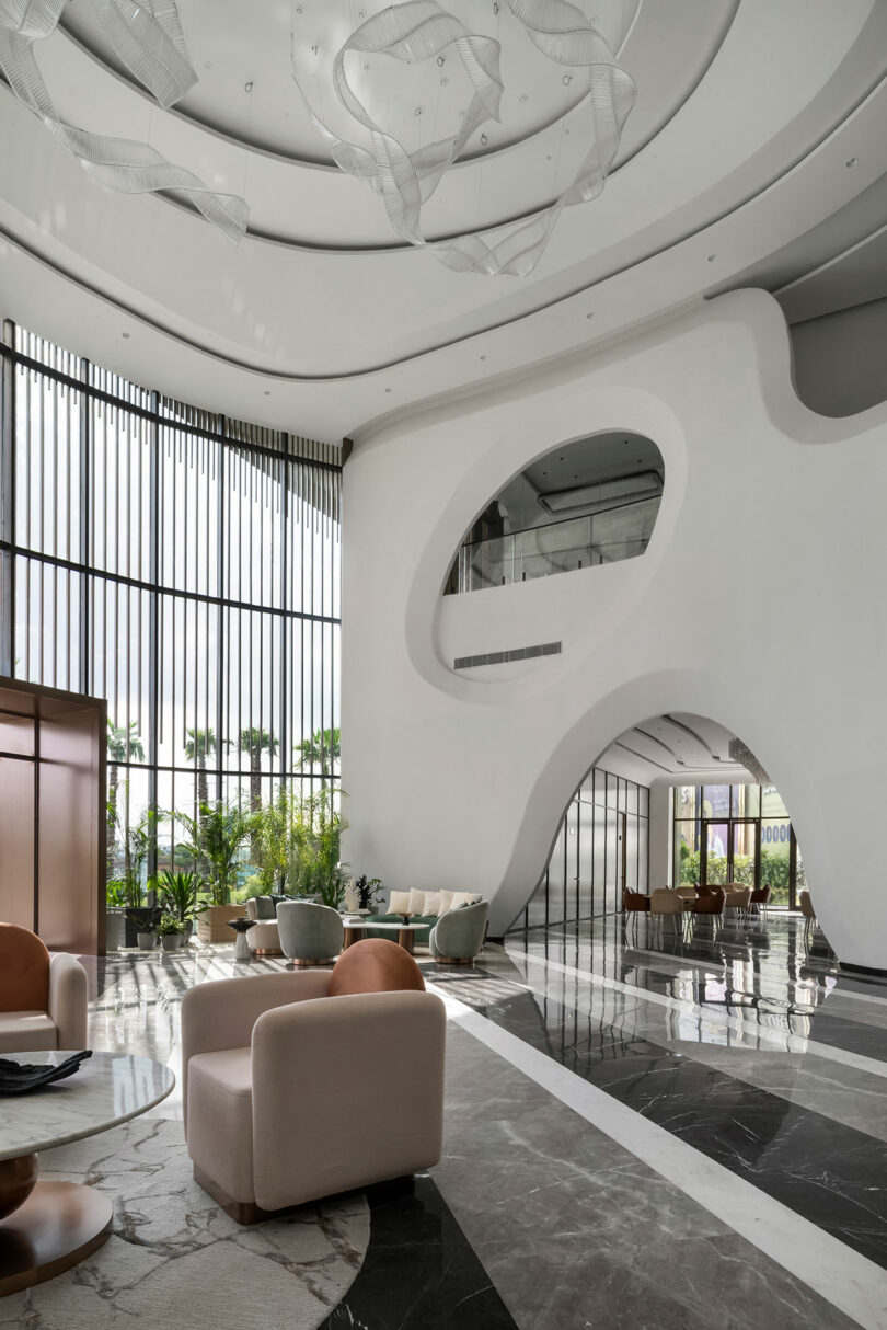 Modern lobby with high ceilings, sculptural white walls, large windows, and contemporary furniture creates a confluence of style and comfort, enhanced by indoor plants and marble flooring in a spacious, well-lit setting.