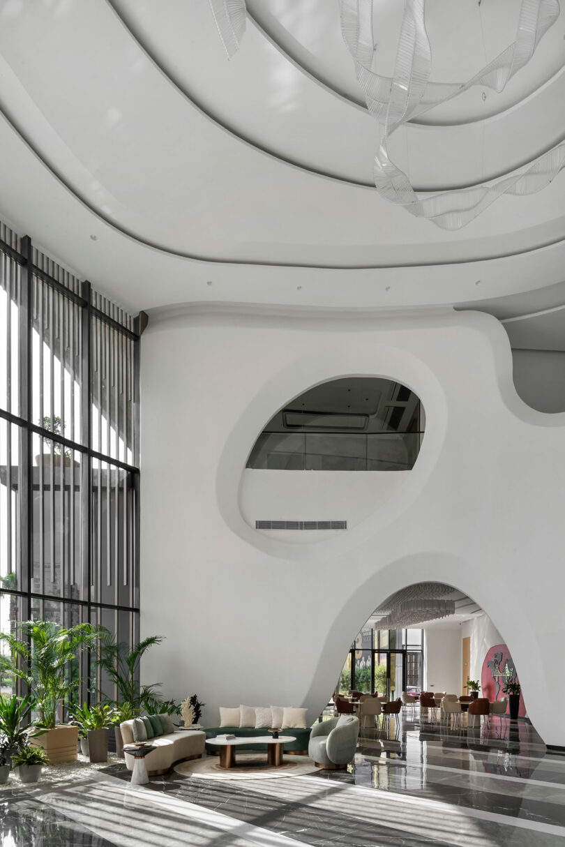 Modern hotel lobby with high ceilings, curved white walls, and large windows creates a confluence of natural light and shadow. Indoor plants and contemporary seating complete the inviting space.