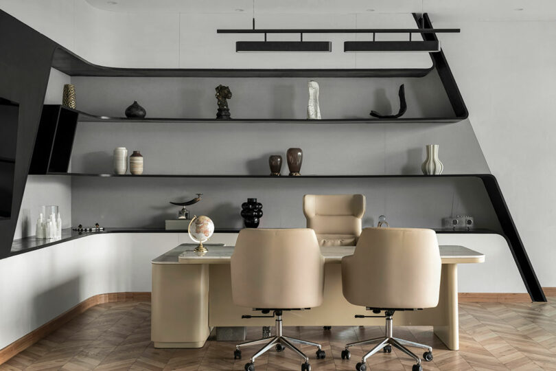 Modern office with two beige chairs, a desk, and curved wall shelves showcasing a confluence of vases, sculptures, and decorative objects against a neutral background.