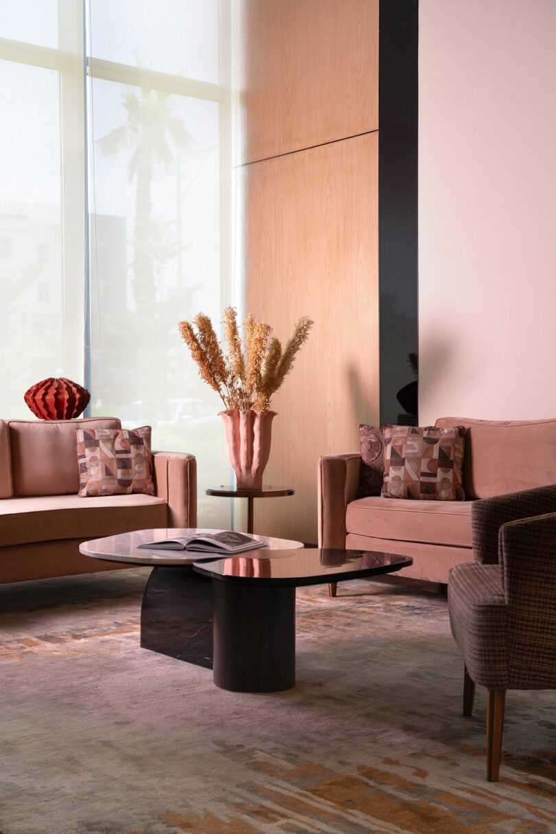 Modern living room with two sofas, patterned cushions, a round coffee table with an open book, and a tall vase with dried flowers near large windows creates a confluence of comfort and contemporary style.