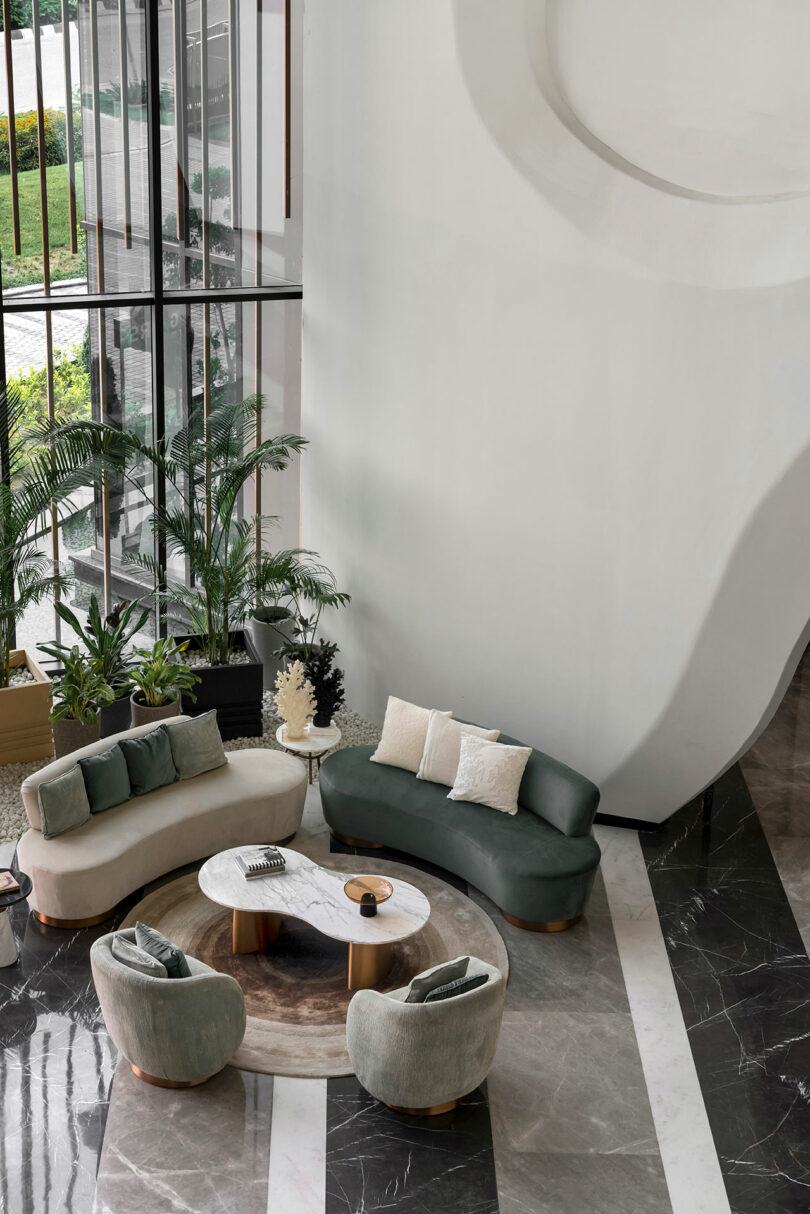 Modern lobby seating area where two curved sofas and armchairs form a confluence around a round coffee table, enhanced by indoor plants and large floor-to-ceiling windows that fill the space with natural light.