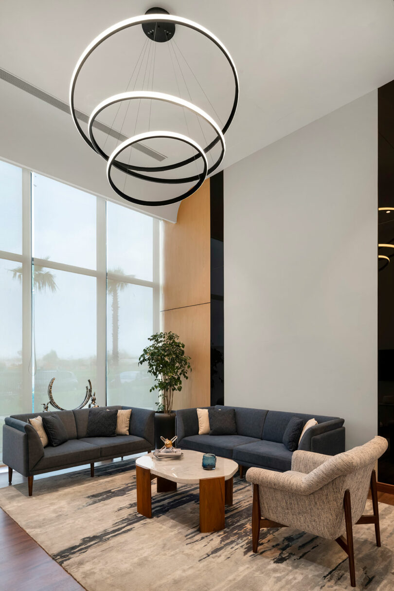Modern living room with large windows where a dark gray sectional sofa and beige armchair form a confluence around a round coffee table, complemented by a geometric rug, potted plant, and circular ceiling light fixture.