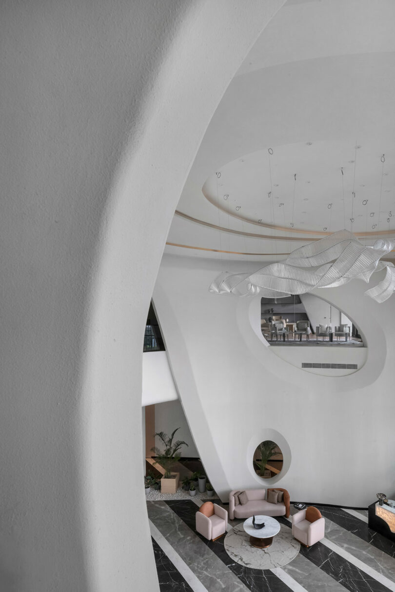 A modern lobby where curved white walls and pink armchairs create a confluence of style, anchored by a round coffee table and sculptural ceiling light. Large circular openings reveal an inviting upper seating area.