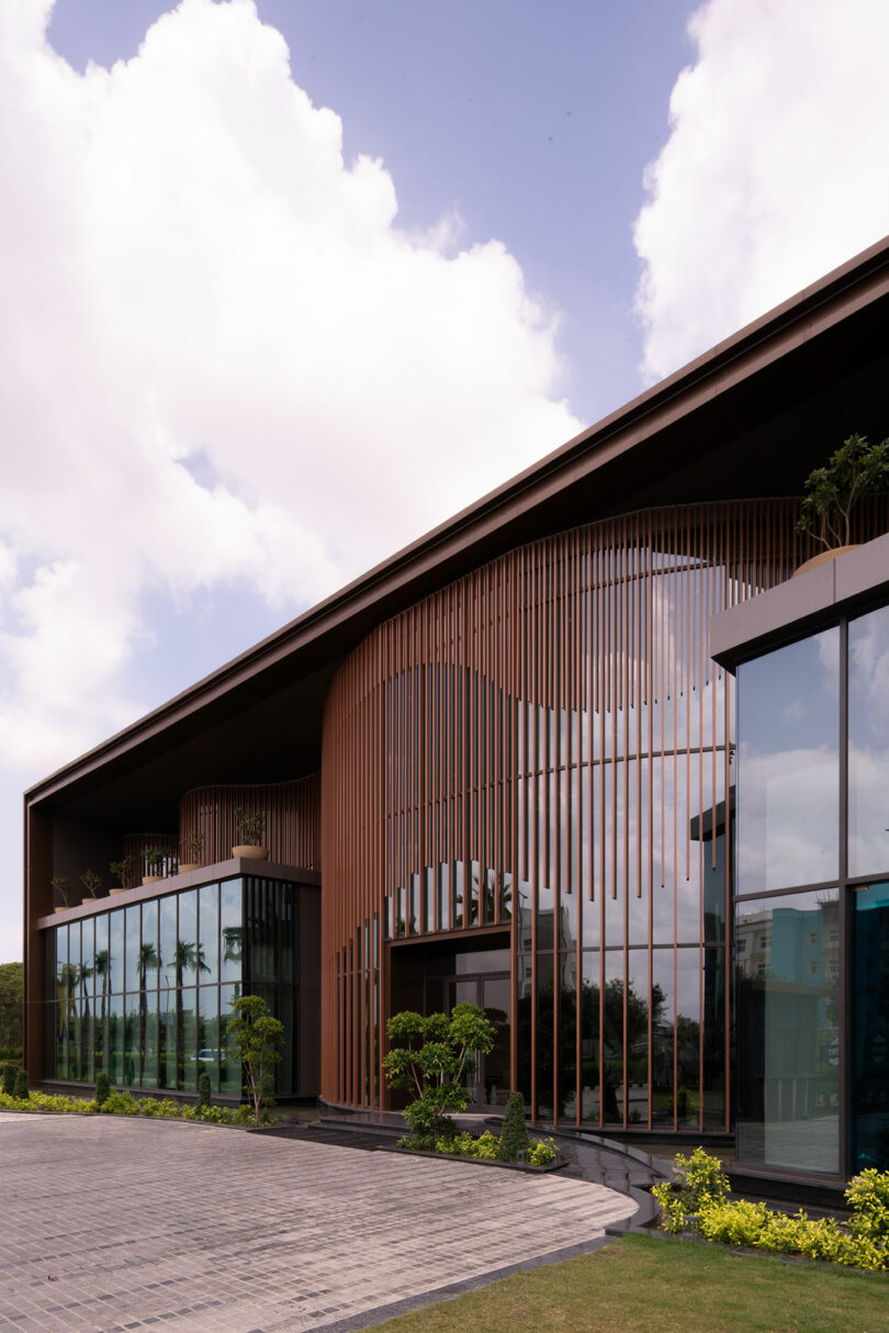 Modern building with large glass windows and vertical wooden slats on the facade, where a confluence of architecture and nature is showcased amid landscaped greenery under a partly cloudy sky.