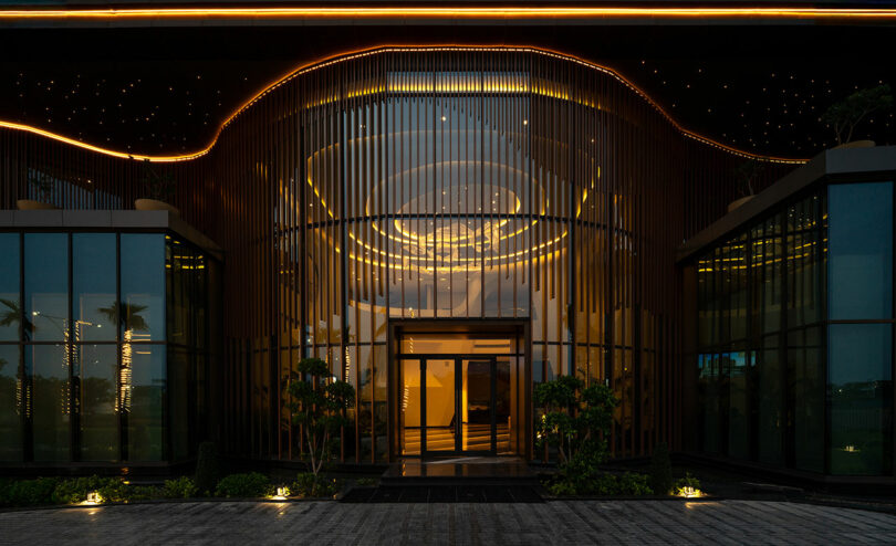 Modern building entrance at night with a curved glass facade, vertical metal slats, illuminated ceiling, and decorative exterior lighting creating a striking confluence of contemporary design elements.