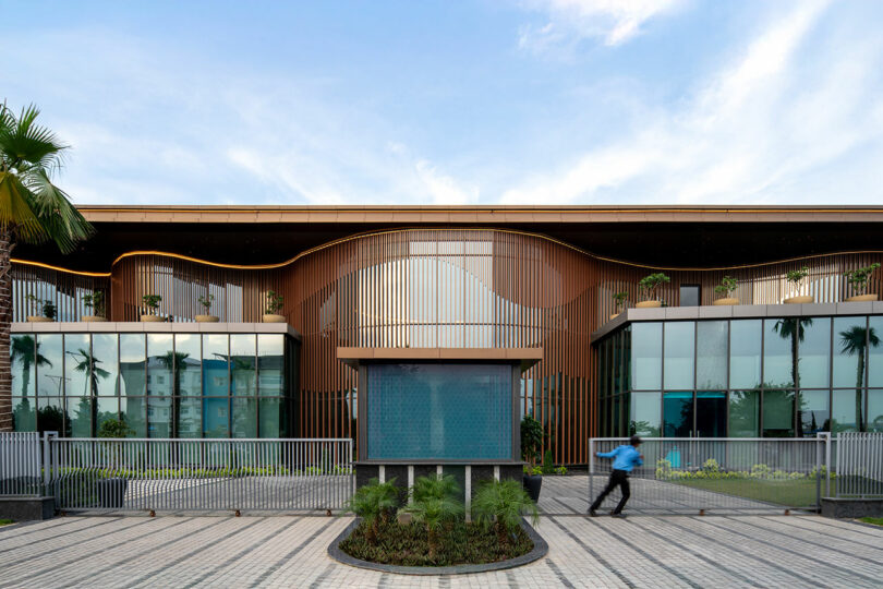 A modern building with large glass windows, vertical wooden slats, and a central water feature creates a striking confluence of design; a person walks past in the foreground under a partly cloudy sky.