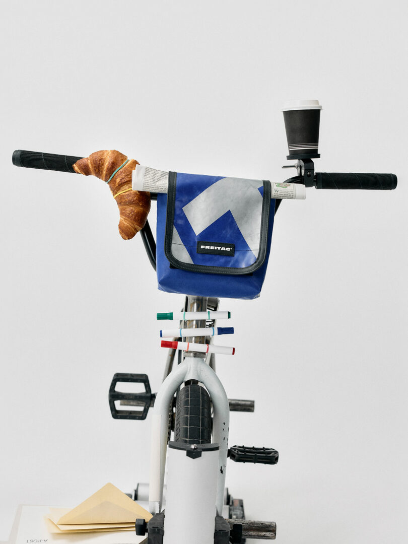 Stationary bike with a blue bag, a croissant on the left handlebar, and a coffee cup on the right handlebar, against a plain white background.