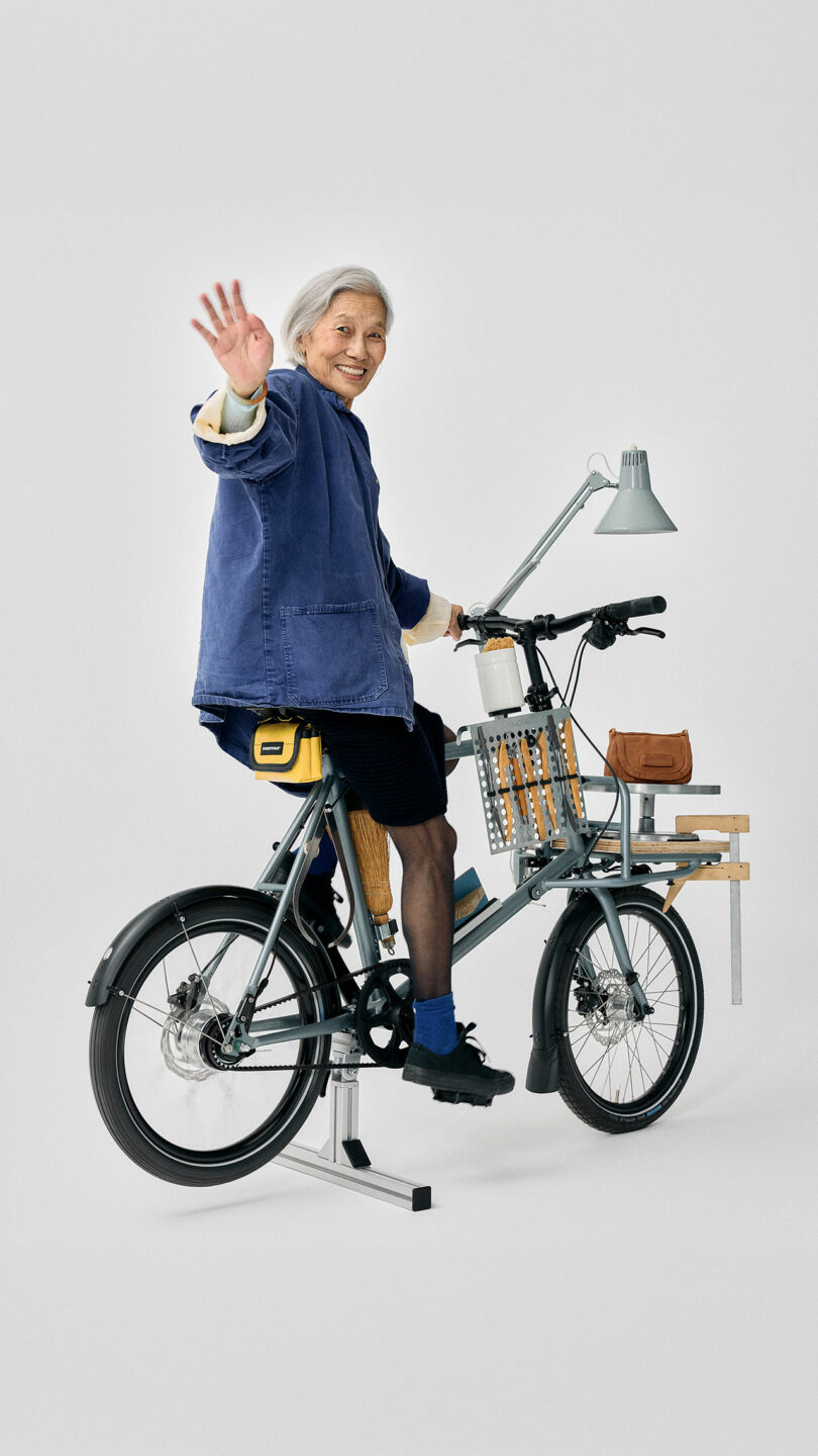 An older woman waves while sitting on a stationary bicycle equipped with a desk, lamp, and small shelves, against a plain white background.