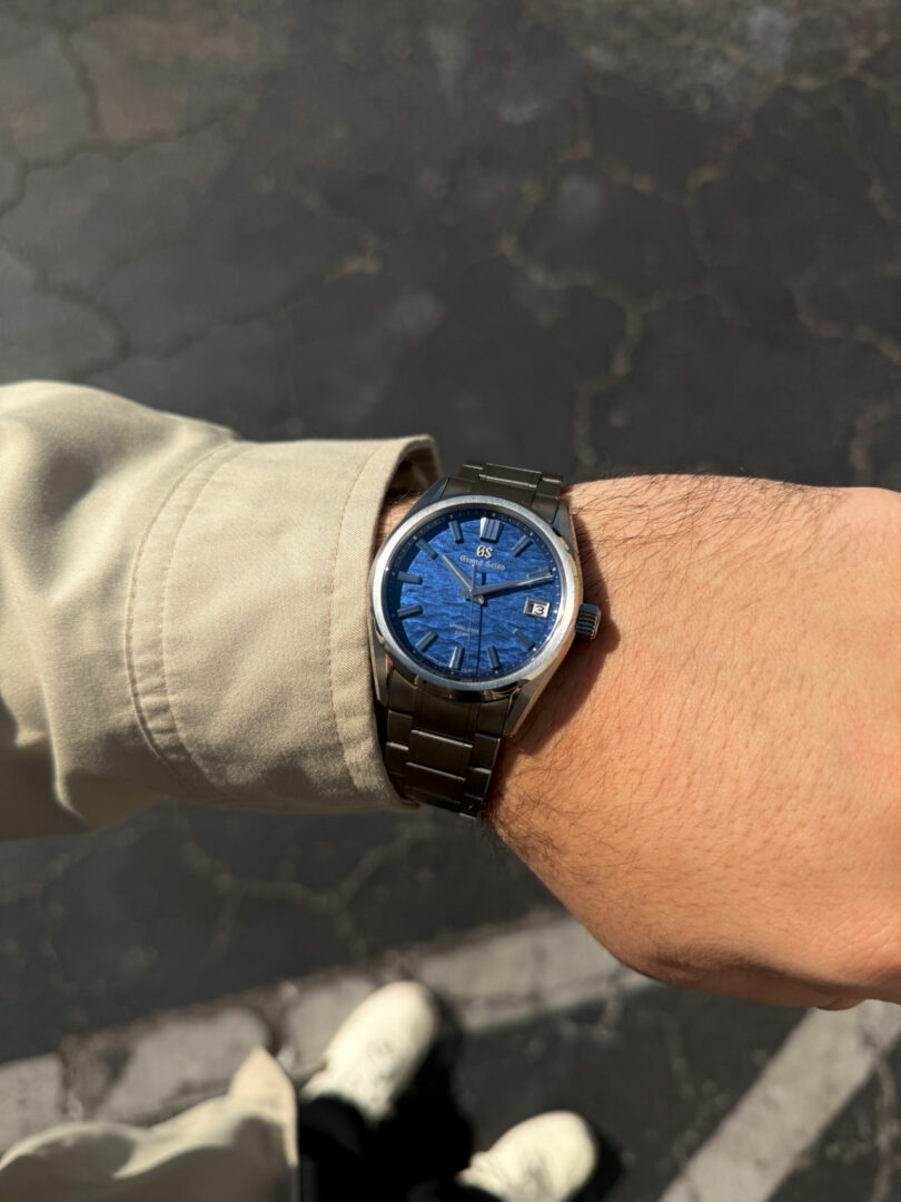 A person wearing a beige sleeve and a black metal Friday Five wristwatch with a blue face stands on a cracked asphalt surface, white shoes visible below.