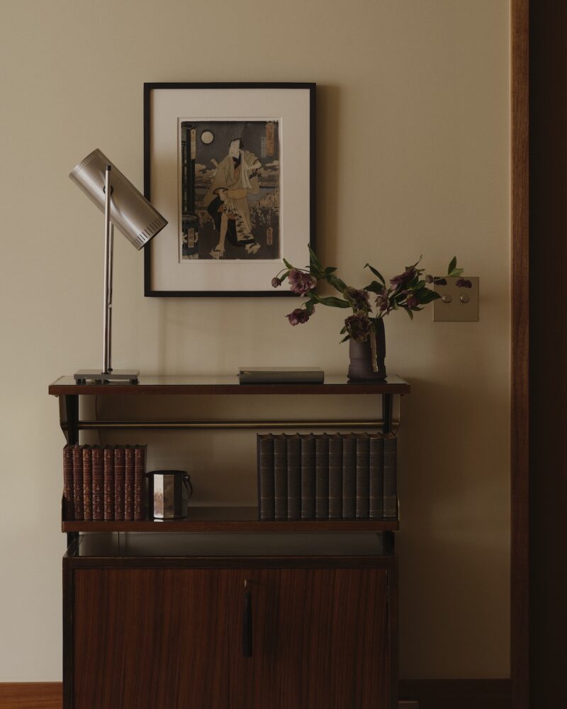A wooden cabinet with shelves holding books, a table lamp, a vase with flowers, and framed art on a beige wall.
