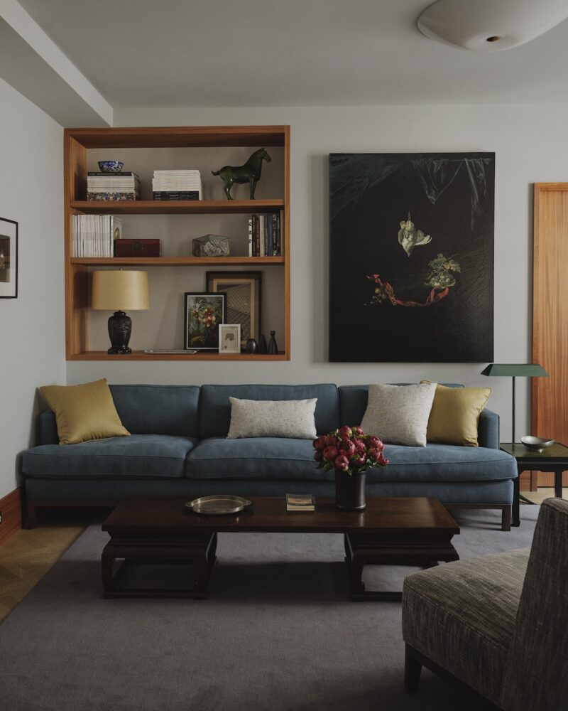 A modern living room with a blue sofa, yellow and white pillows, a wooden coffee table with a vase of flowers, built-in shelves, and a large dark painting on the wall.