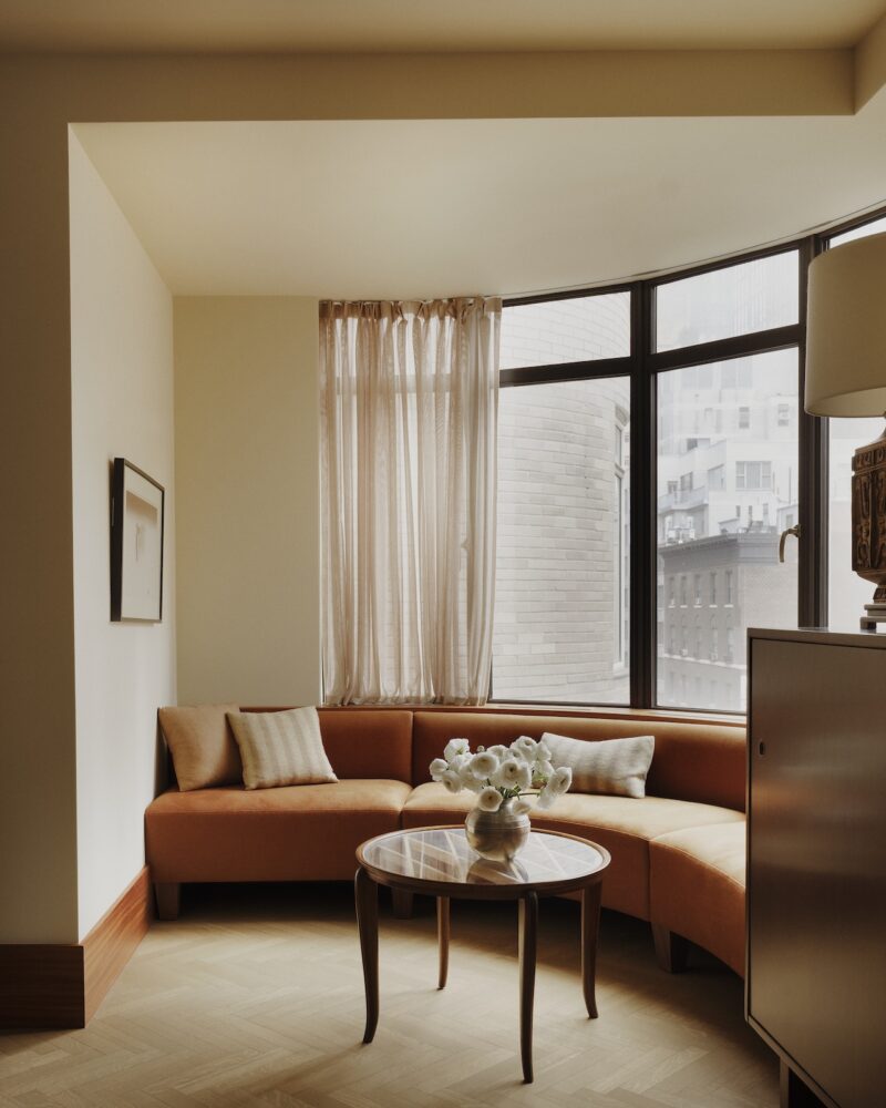 A curved orange sofa with beige cushions sits by a large window in a modern room, with a round coffee table holding a vase of white flowers in front.