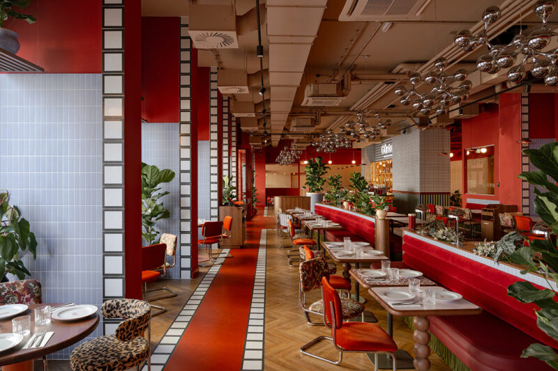 A modern restaurant interior by Atelier OLK with red walls, patterned floors, red upholstered seating, set tables, plants, and silver globe pendant lights.