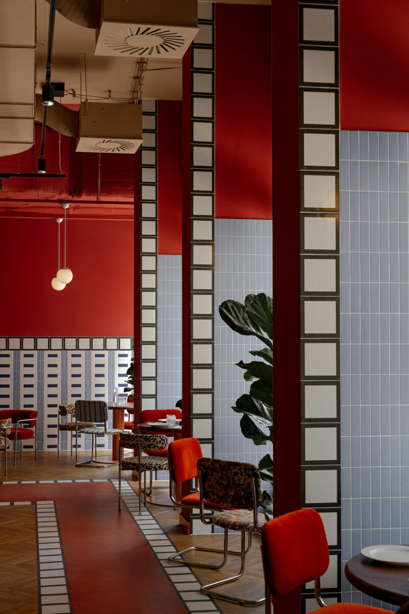 A modern restaurant interior by Atelier OLK features red walls, blue tiled columns, geometric patterns, and tables with red and floral chairs. Large leafy plants add vibrant greenery to the inviting space.