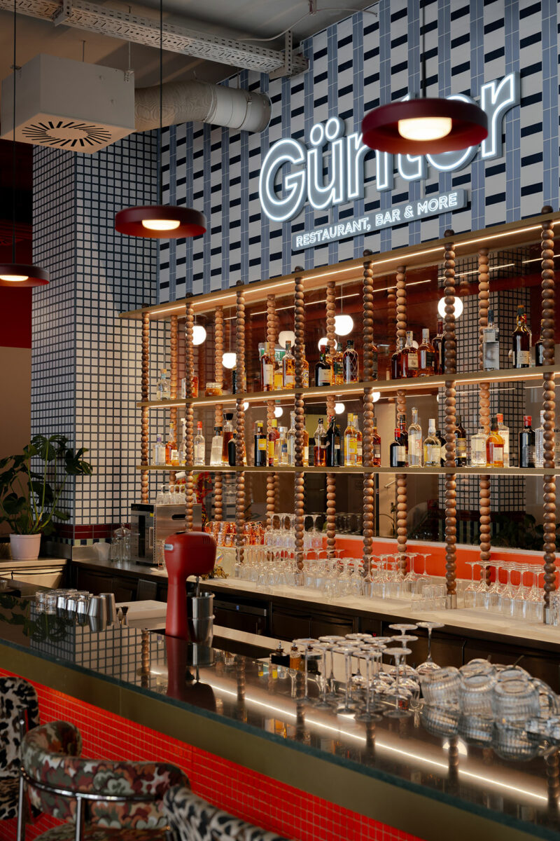 A modern bar by Atelier OLK features a tiled wall, an illuminated "Günter" sign, shelves stocked with bottles and glassware, red pendant lights, and a countertop adorned with cups and a red coffee machine.