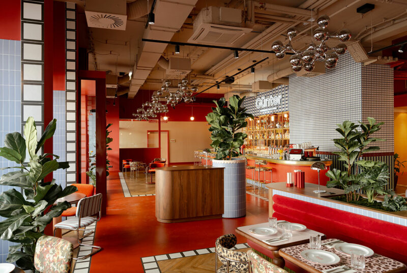 Modern restaurant interior by Atelier OLK featuring red floors, tiled walls, lush green plants, and contemporary lighting. Tables are set for dining, with a stylish bar area visible in the background.