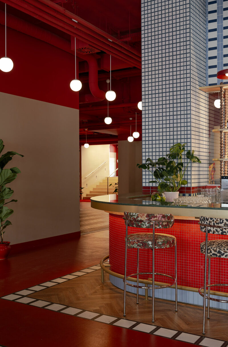 A modern bar by Atelier OLK with red tile, animal print barstools, globe pendant lights, a tiled column, lush plants, and wood flooring in a vibrant, colorful interior.