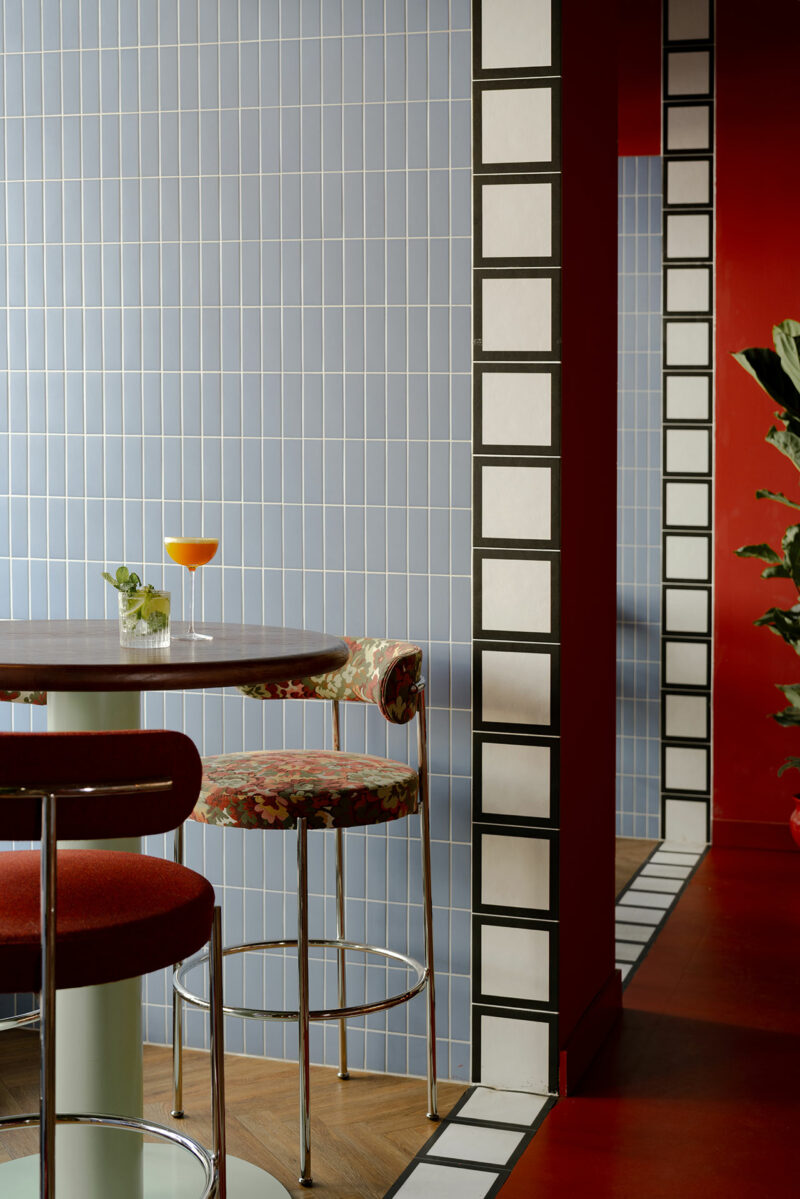 A cocktail and a glass of water sit on a round table next to two patterned bar stools; the room, designed by Atelier OLK, features blue tiled walls and bold red and white accents.