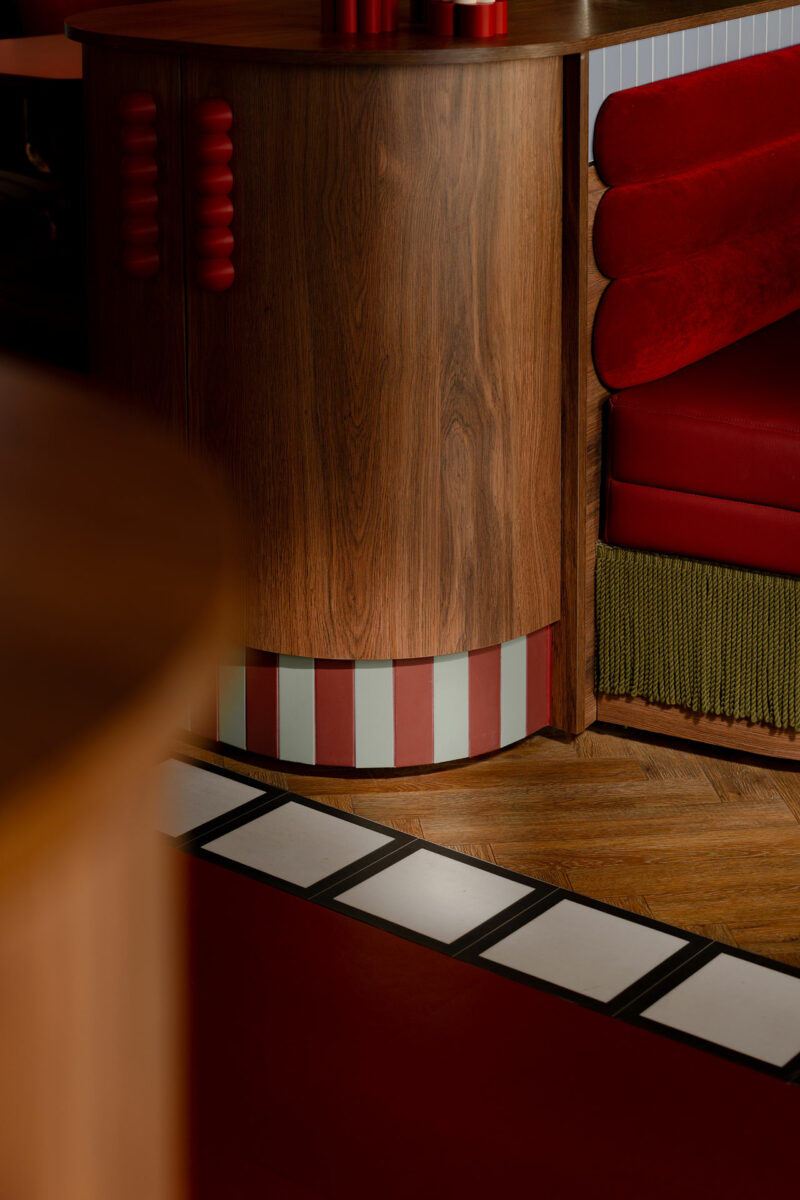 A close-up of a wood-paneled corner by Atelier OLK, featuring red and white vertical stripes at the base, red upholstered seating, and a floor with wood, red, and black-and-white tile patterns.