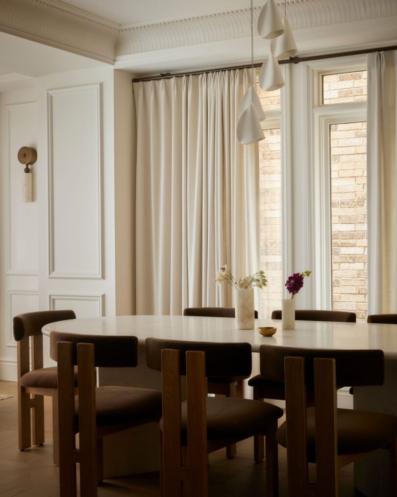 Modern dining room with a round table, dark cushioned wooden chairs, vases with flowers, beige curtains, pendant lights, and a window showing a brick wall outside.