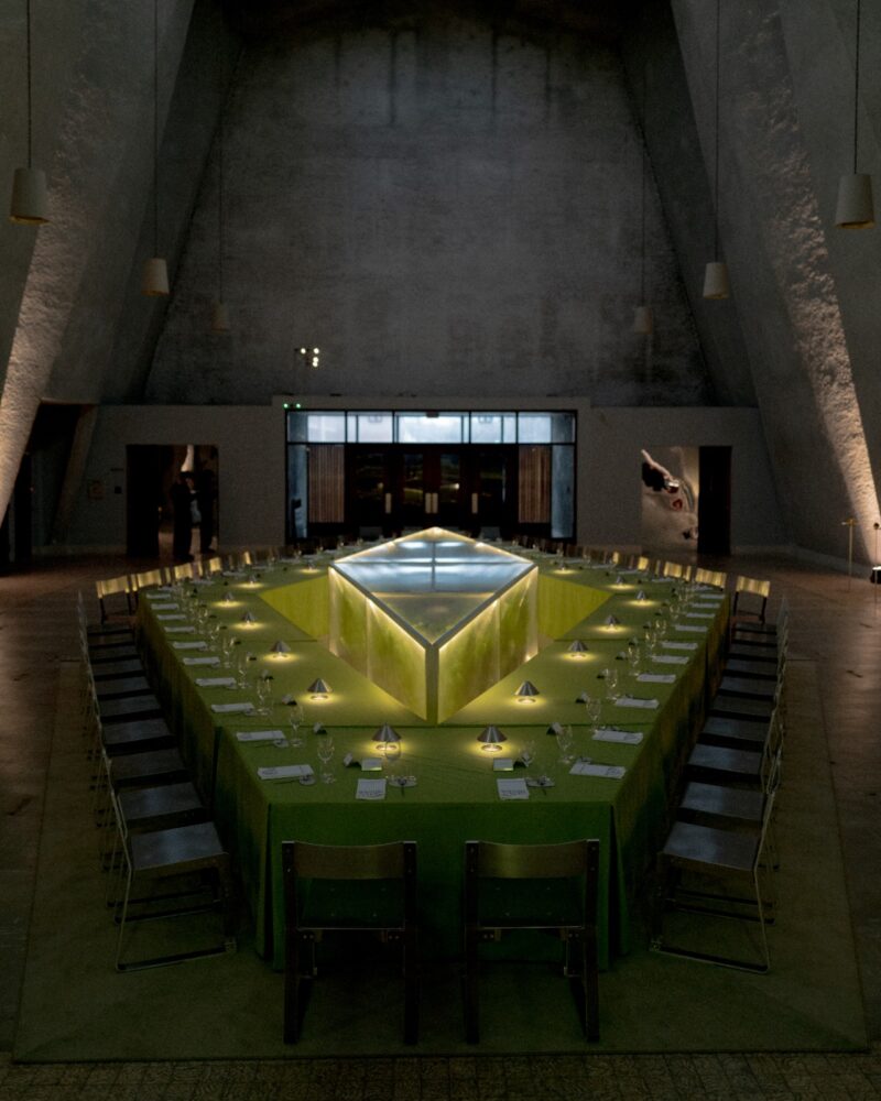 A large, triangular dining table set for a formal meal is arranged in a dimly lit, modern space with high concrete walls, hanging lights, and a Menu Extra to enhance the dining experience.