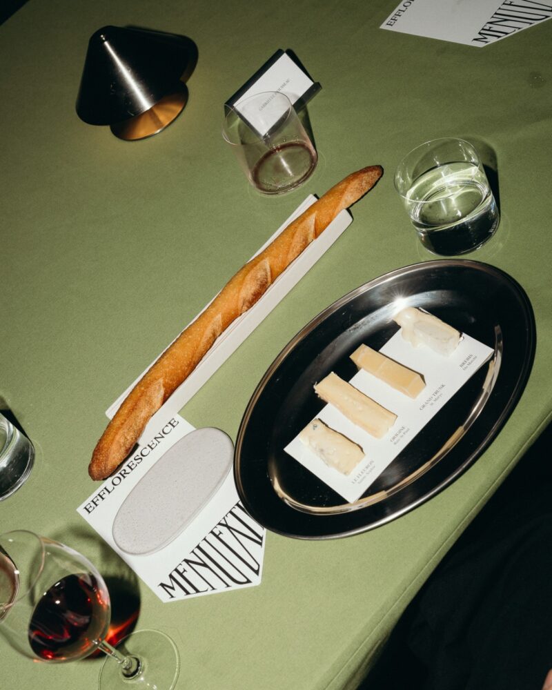 A table set with a baguette, a plate of four labeled cheeses, a glass of red wine, a glass of water, and Menu Extra options atop a green tablecloth.