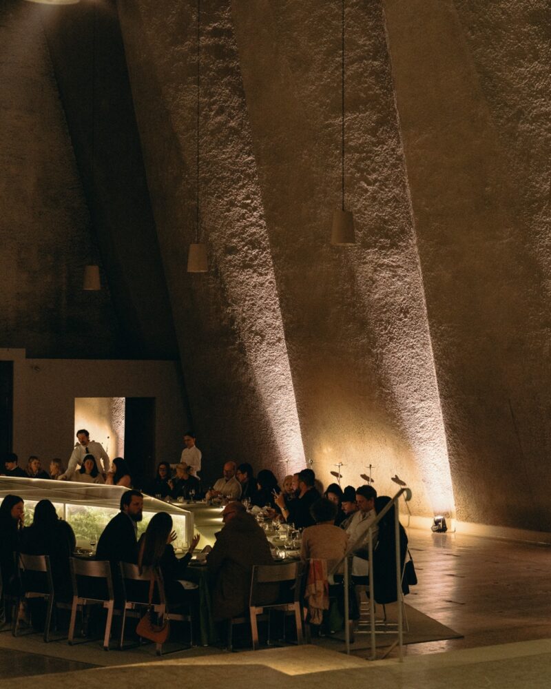 A group of people sit around a curved bar in a dimly lit, modern space with tall textured walls and hanging pendant lights, browsing the Menu Extra for unique offerings.