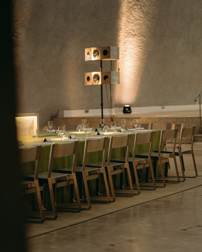 Long dining table set with glassware and napkins, surrounded by wooden chairs in a spacious, modern room with industrial lighting, textured walls, and a Menu Extra for special selections.