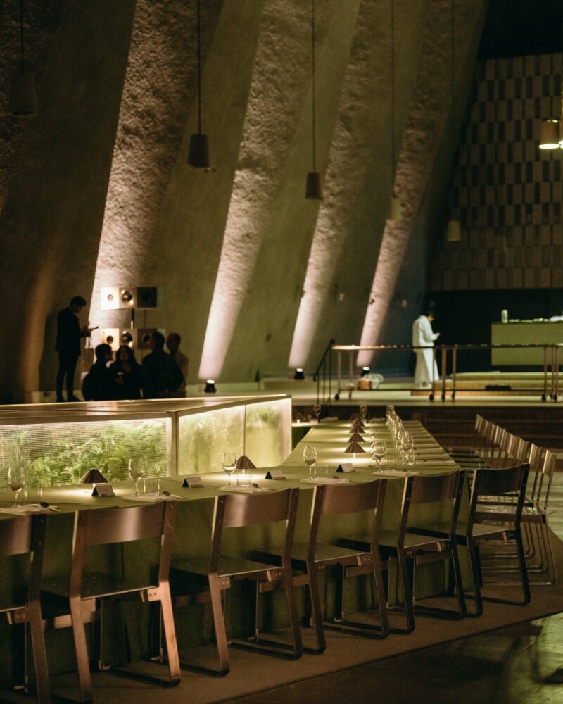 A modern dining area with a long table set with wine glasses, plates, and stylish Menu Extra touches, illuminated by soft lighting, with people standing and sitting in the background.
