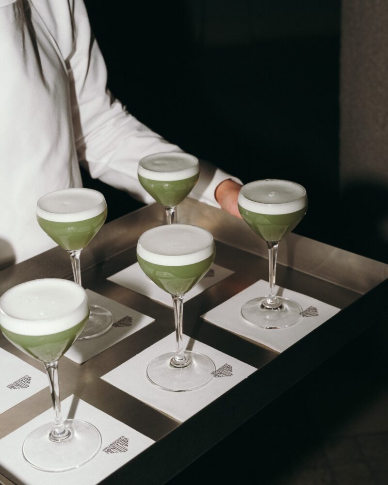 A server holding a tray with five coupe glasses filled with a green cocktail topped with white foam, each placed on a napkin—an eye-catching menu extra for your next event.