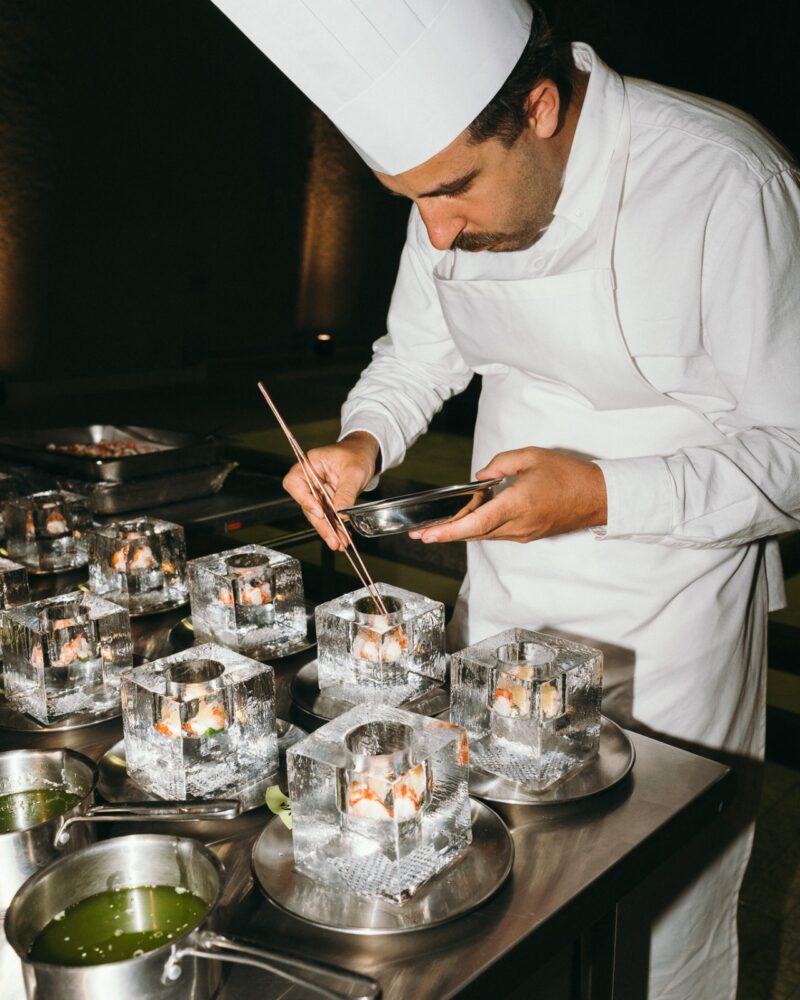 A chef in a white uniform and hat is carefully plating food into square ice blocks on a stainless steel table, preparing a unique Menu Extra for guests.