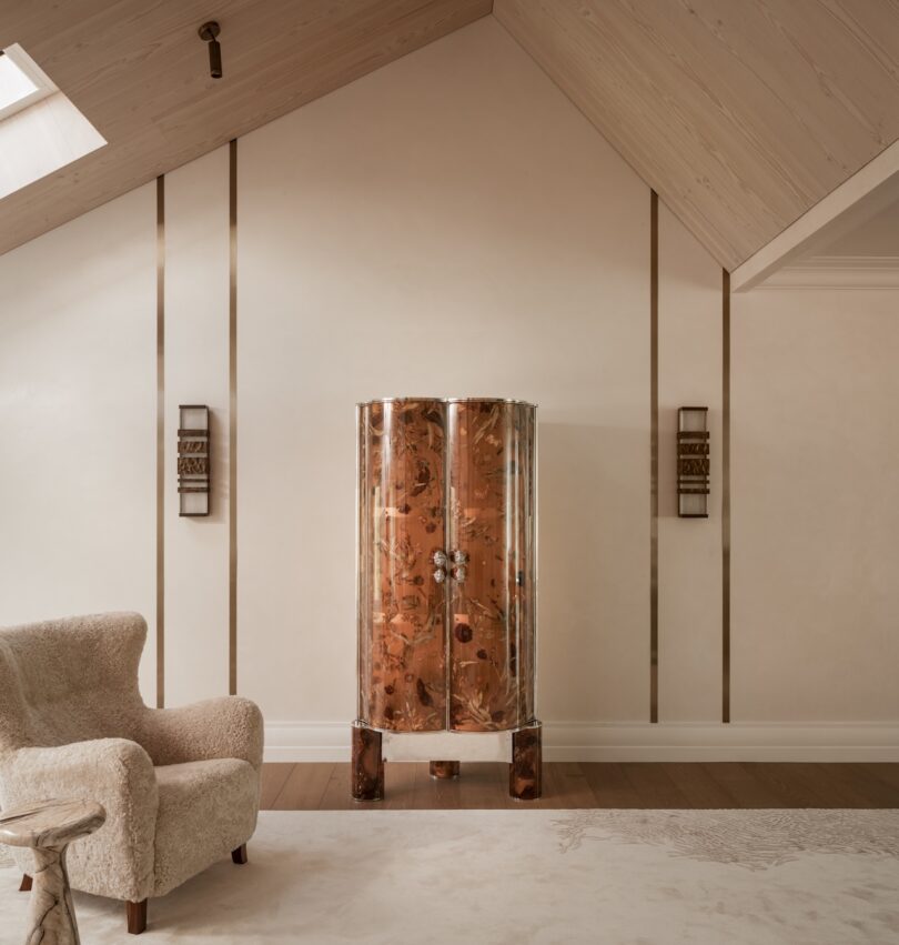 A plush beige armchair sits next to a tall, glossy copper cabinet against a cream-toned wall with gold trim in a modern, minimalist room with slanted ceiling.