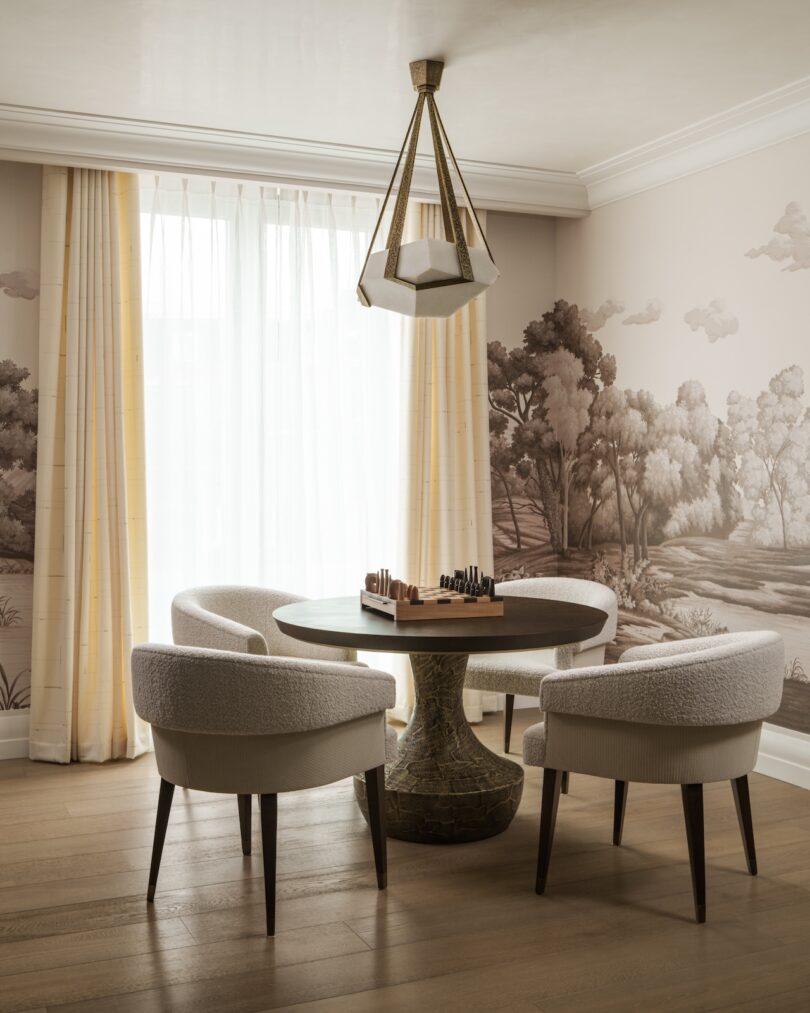 Round table with a chess set surrounded by four upholstered chairs in a room with light curtains, wooden floor, and nature-themed wall mural. Geometric pendant light overhead.