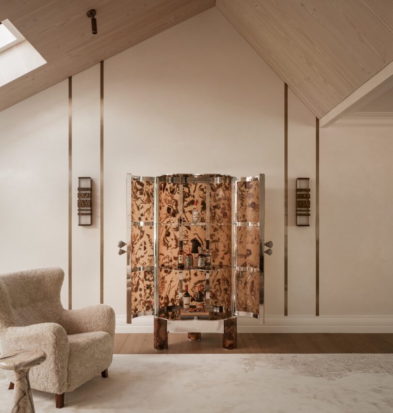 A decorative, metallic cabinet with open doors stands against a beige wall in a room with a sloped ceiling, next to a textured armchair and a small wooden side table.
