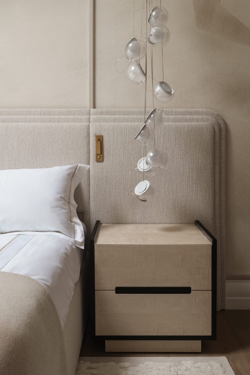 A modern bedside table with two drawers stands next to a bed with white linens; several glass teacups hang above on transparent strings as decoration.