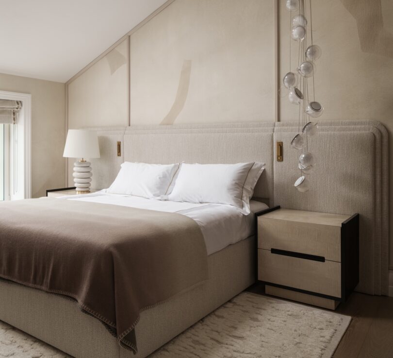 Neutral-toned bedroom with a large upholstered bed, white and beige bedding, two nightstands, a modern lamp, hanging glass decor, and a soft area rug.