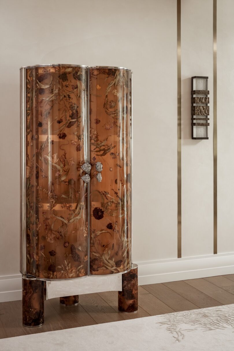 Curved cabinet with a brown floral pattern, chrome edges, and decorative handles stands against a beige wall with metallic vertical accents and a wall sconce.