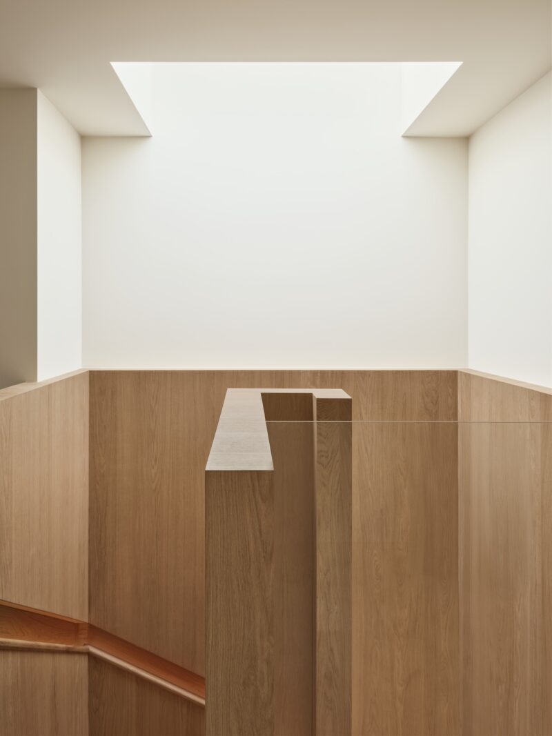 Minimalist interior with light wood paneled staircase and walls, illuminated by a large rectangular skylight in a white ceiling.