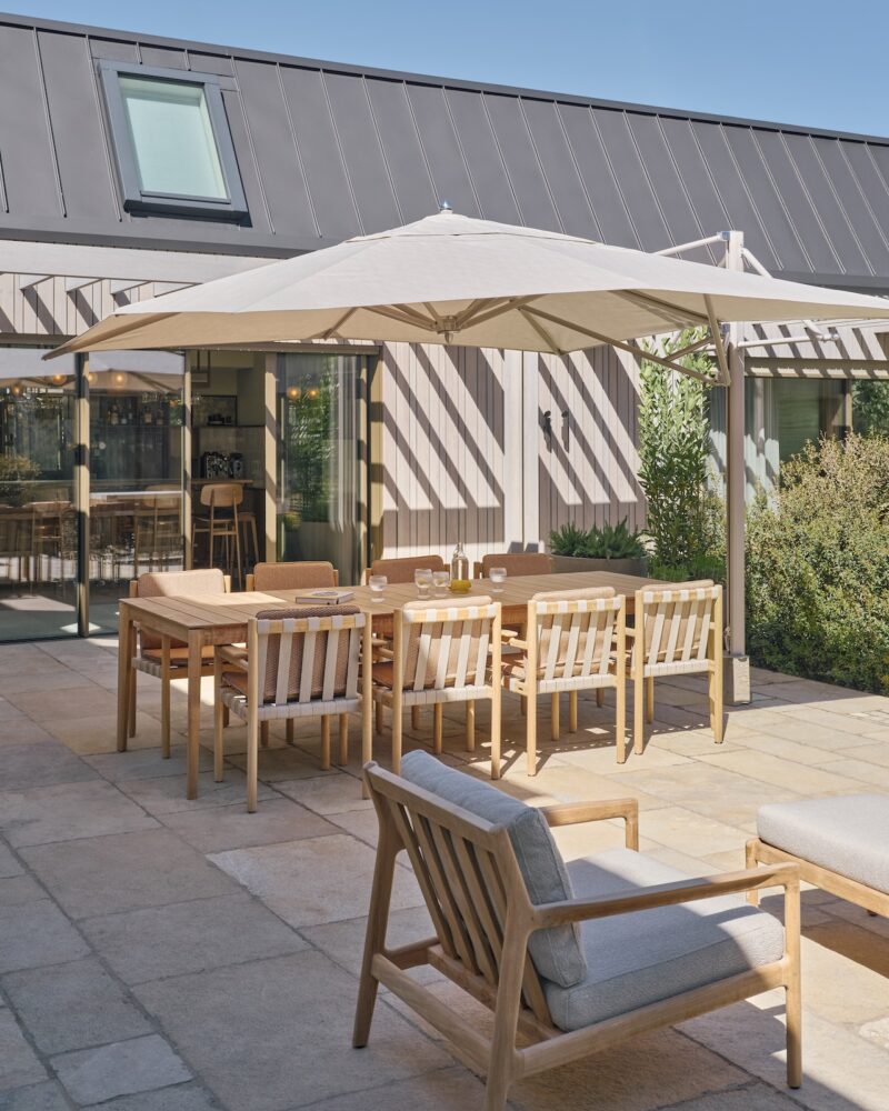 Outdoor patio with wooden dining table and chairs under a large umbrella, set on stone pavers near a modern building with sliding glass doors.