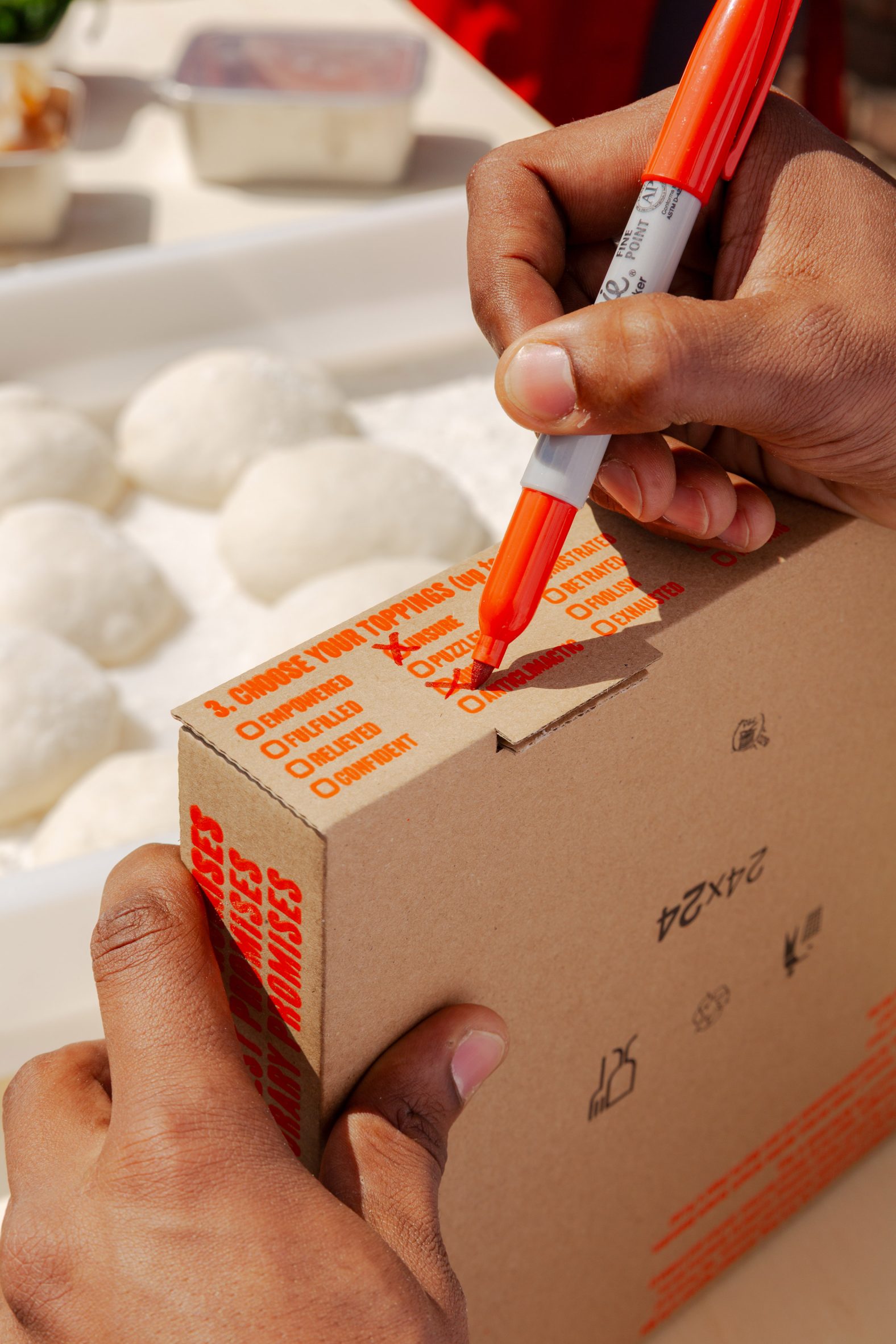 Close-up photo of a person's hands as they fill out a checkbox list on the side of a pizza box with words like empowered, fulfilled and relieved
