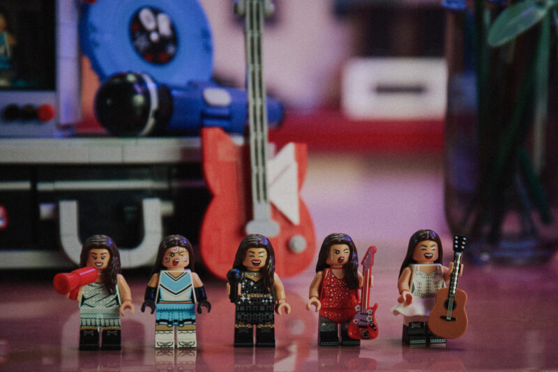 Five LEGO minifigures dressed as Olivia Rodrigo stand in a row, with various musical instruments and equipment in the background.