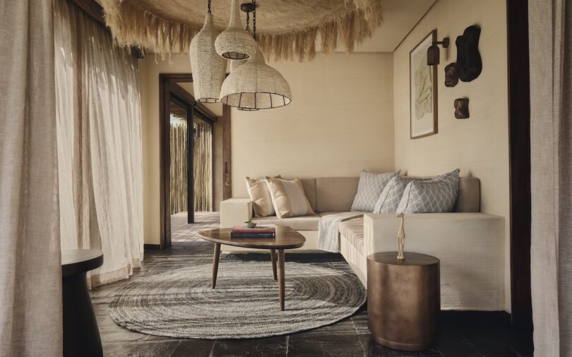 A neutral-toned living room with a sectional sofa, round coffee table, woven rug, and hanging wicker lamps, featuring natural light and minimalist decor.
