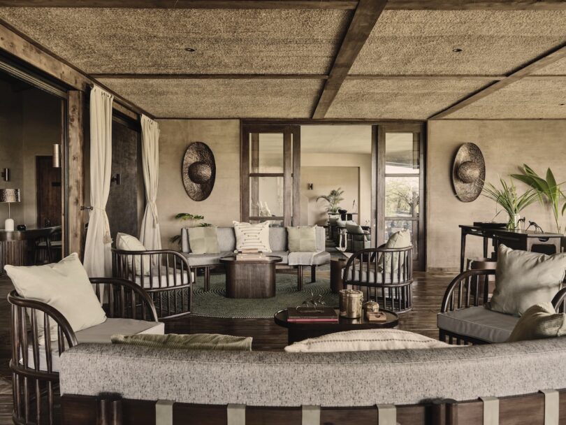 Spacious living room with wooden furniture, neutral cushions, woven decor on walls, glass doors, indoor plants, and a large rug on wooden floor.