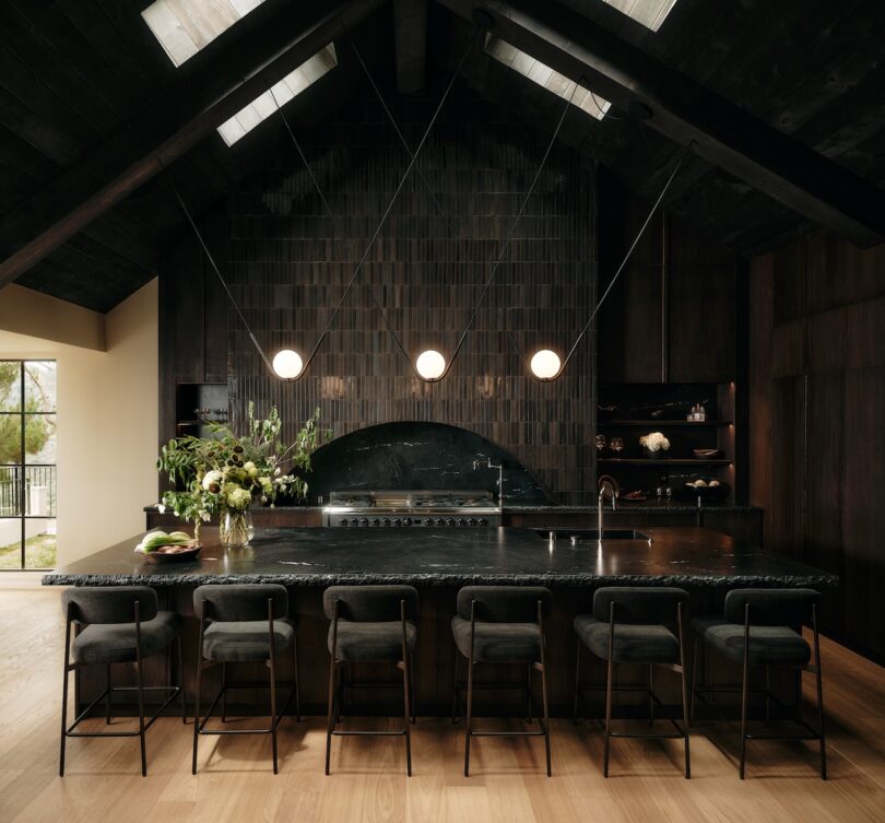 Modern kitchen with a dark, vaulted ceiling, black tile backsplash, large island with six barstools, pendant lights, and wood flooring.