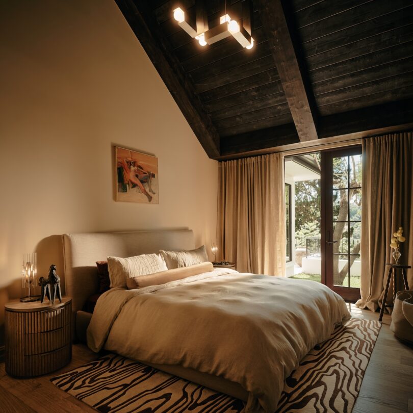 A cozy bedroom with a large bed, beige bedding, wooden ceiling, zebra-pattern rug, side table with candles, wall art, and glass doors opening to a patio with greenery outside.