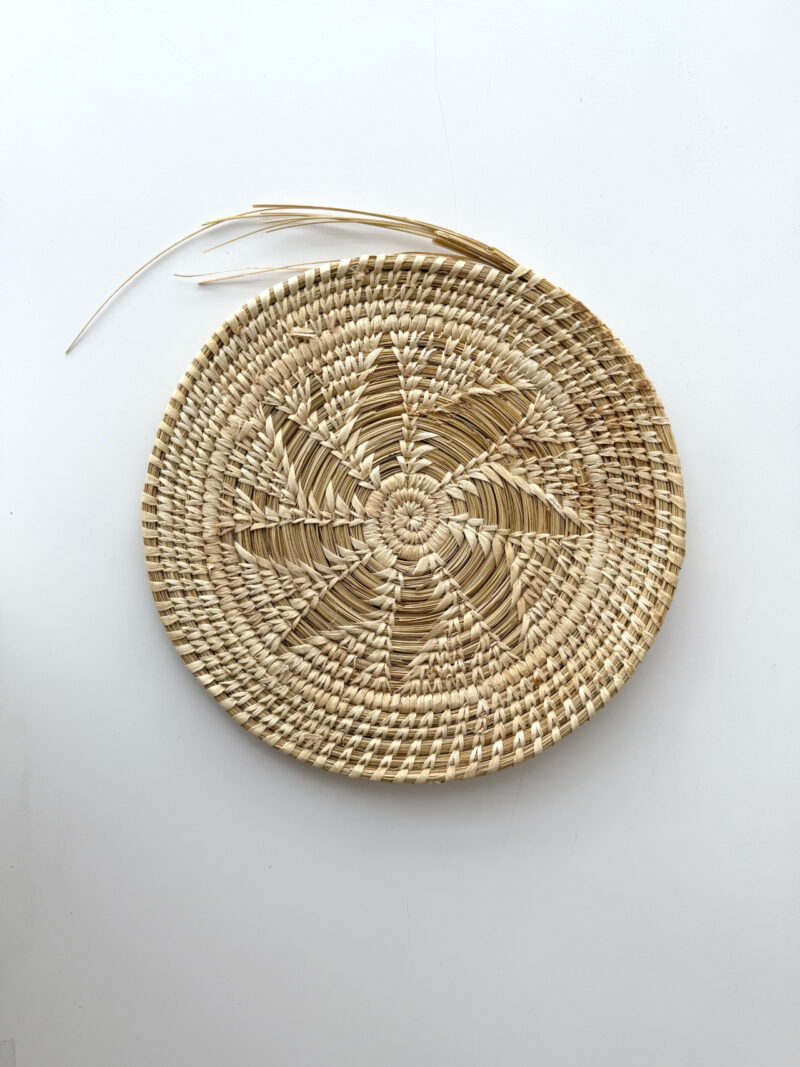 Round woven straw mat with a star pattern in the center, resting on a plain white background; some loose straw strands are visible at the top.